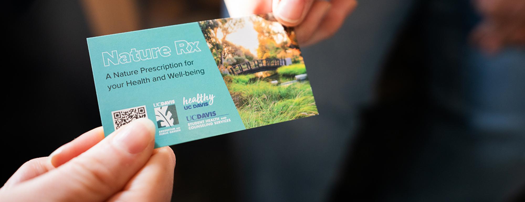 Image of a hand giving another hand a business-type card with a beautiful outdoor image of a bridge in the UC Davis Arboretum and includes the copy:  Nature Rx A Nature Prescription for your Health and Well-being. The card includes a QR code and wordmarks from the participating partners including the UC Davis Arboretum and Public Garden, Healthy UC Davis, and UC Davis Health and Counseling Services.