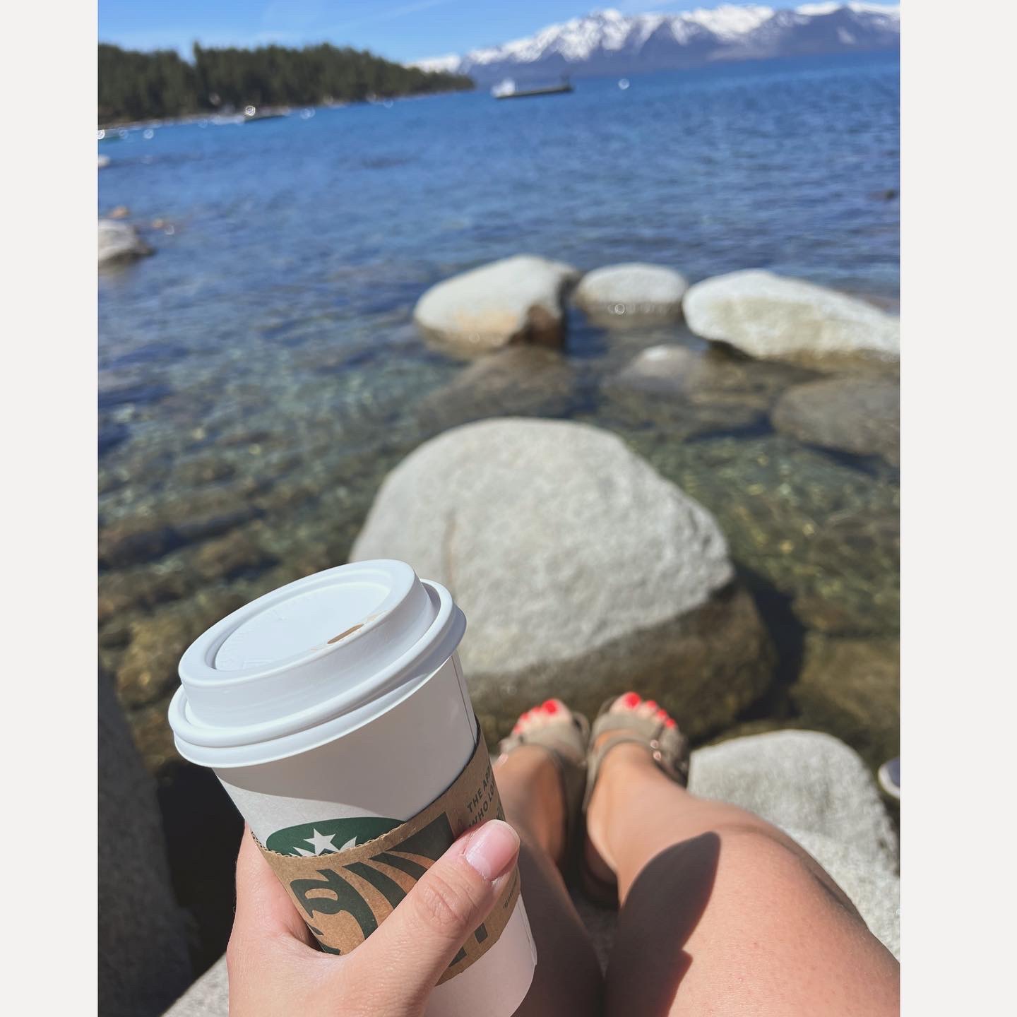 Person sitting on rocks on the coast of a lake, holding a Starbucks cup