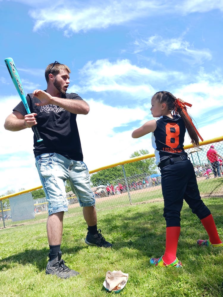 A kid wearing a softball uniform and an adult holding a bat at a baseball field