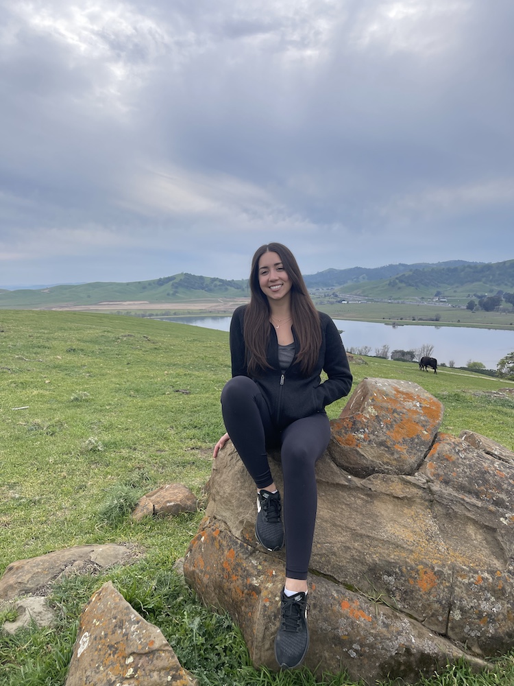 Person sitting on a large rock, with grassy hills and a lake in the background