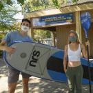 two students posing with a kayak and holding an oar outside of the Outdoor Adventures office