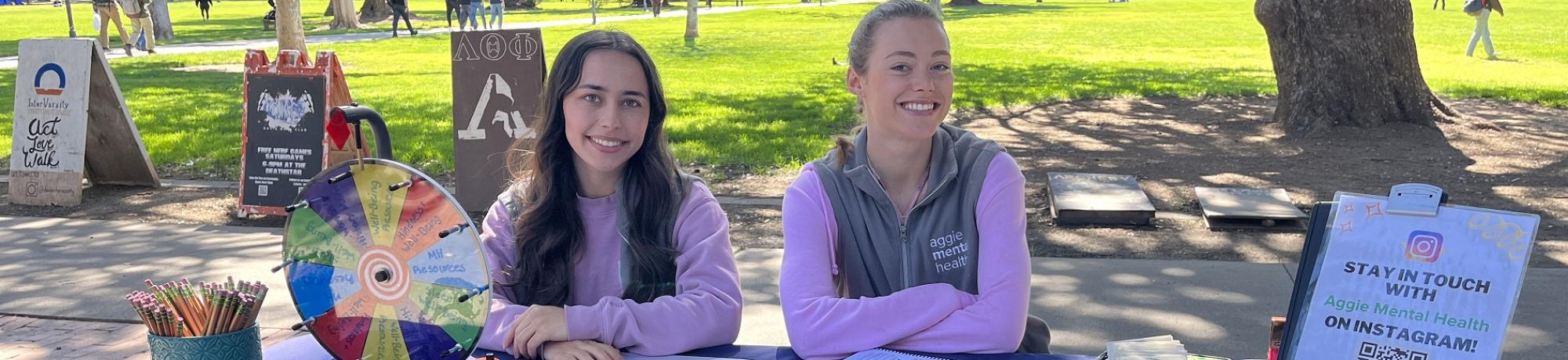 two students sitting at a table with a tablecloth saying "aggie mental health"
